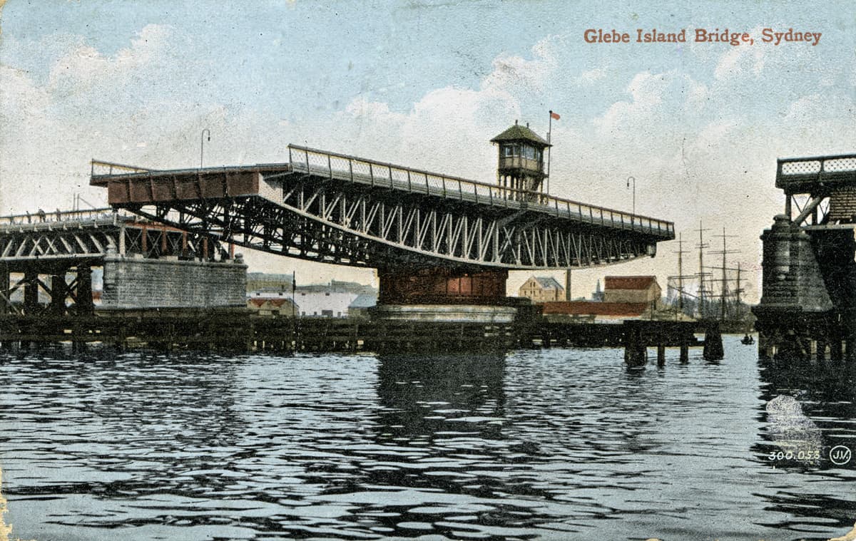 Coloured postcard view of the bridge, swung open to allow shipping to pass through. On reverse personal message about weather and sightseeing, "like Sydney very much". 1915. Image courtesy of City of Sydney Archives
